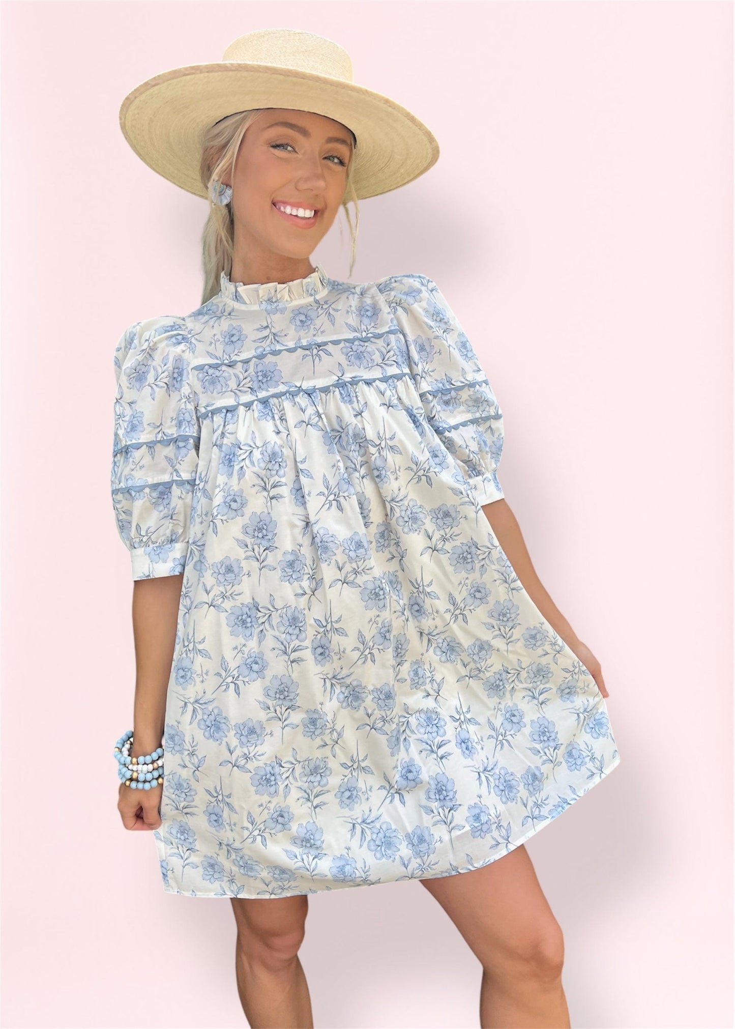Model wearing the floral babydoll mini dress with puff sleeves and mock neckline, showcasing the swing silhouette and delicate blue floral print on a cream background.