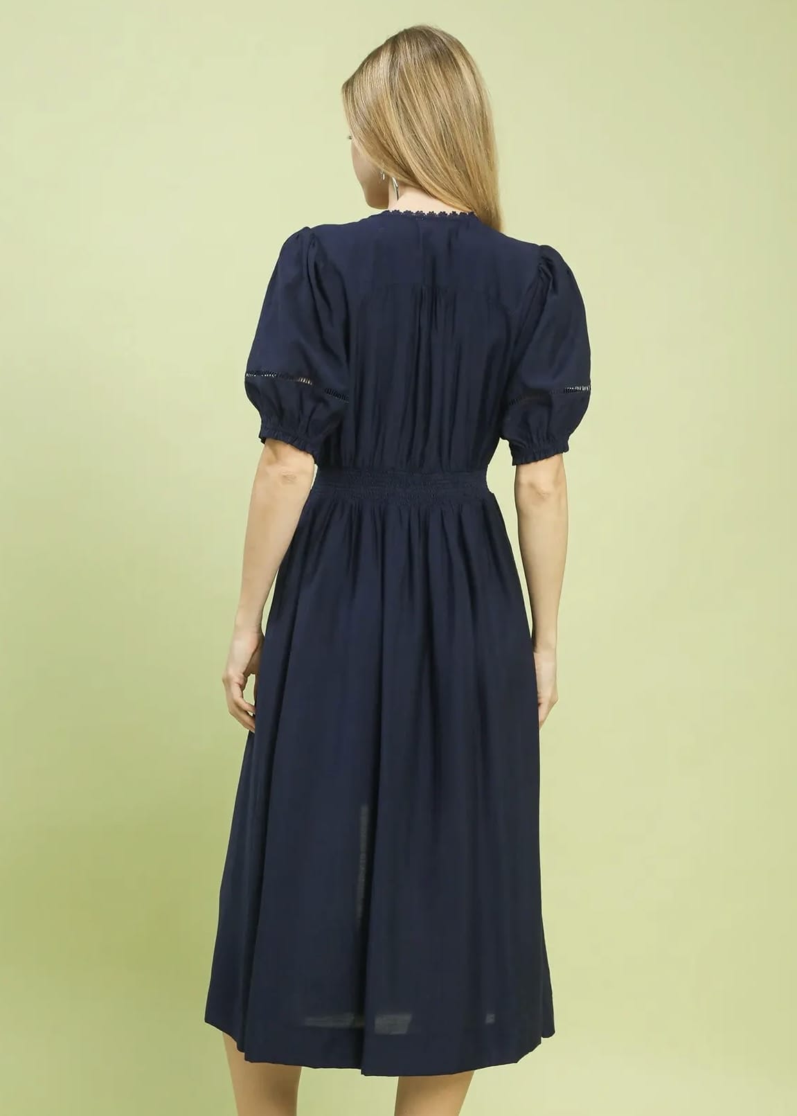 A woman viewed from the back wearing a navy blue midi dress with short puff sleeves, a gathered waist, and a pleated back.