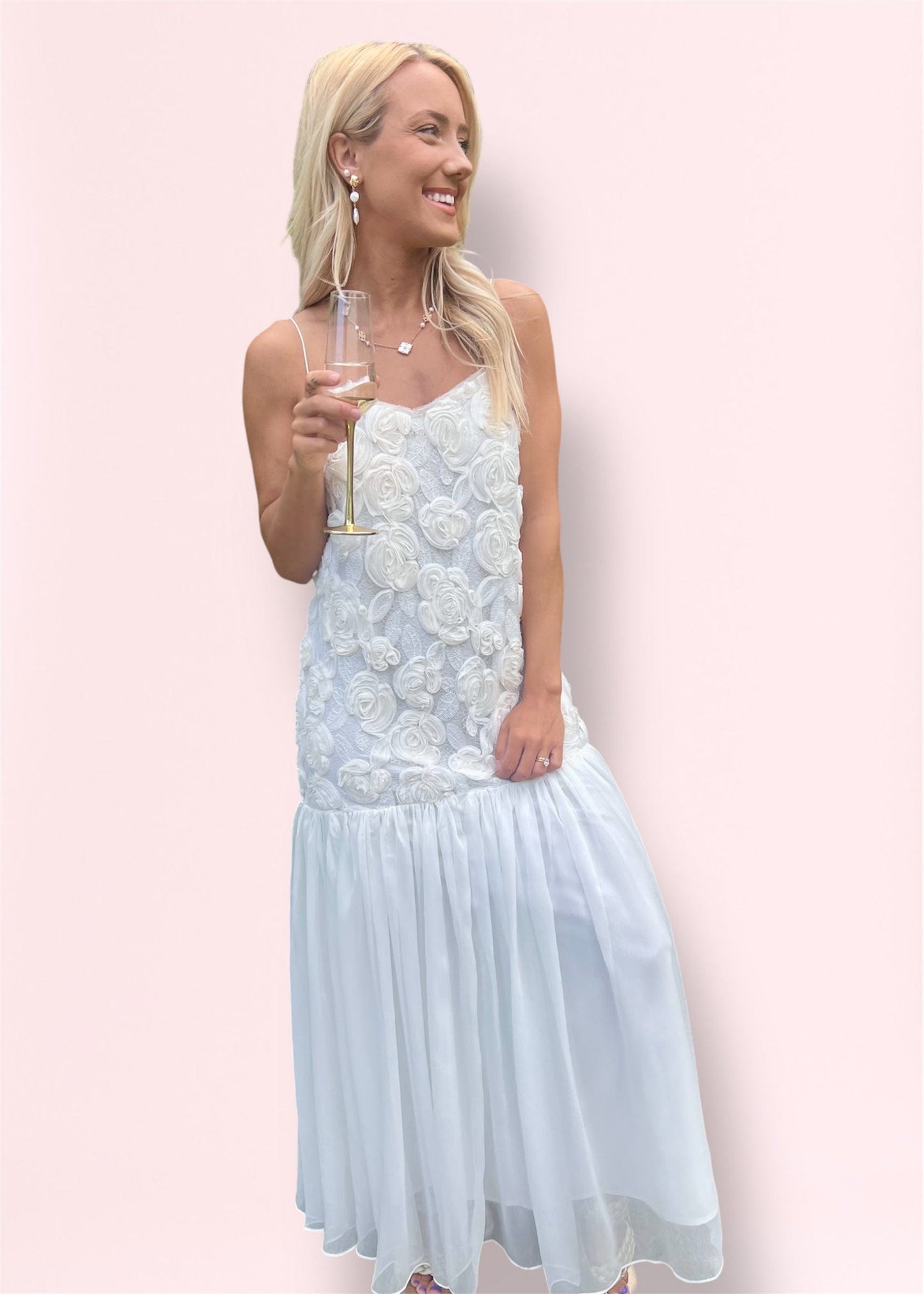 model holding champagne glass in romantic white floral maxi dress with rosette bodice and sheer flowy skirt.