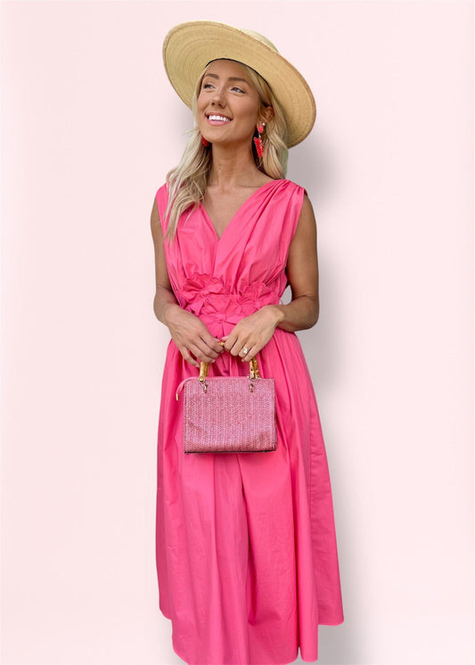 Model wearing the Ilona Coral Midi Dress with deep V-neckline and floral waistband detail, styled with a wide-brim straw hat, statement coral earrings, and pink woven handbag—perfect for spring and summer occasions.