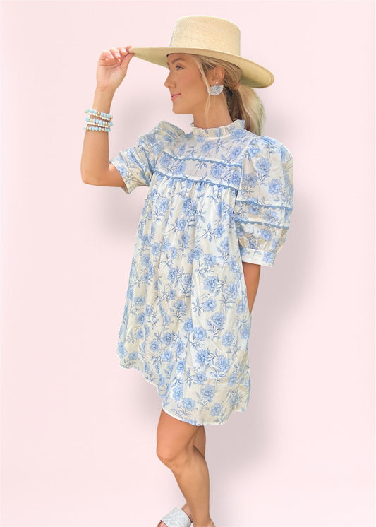 Side view of model wearing a floral babydoll mini dress with puff sleeves, mock neckline, and a blue-on-white print, accessorized with a straw hat and stackable bracelets.