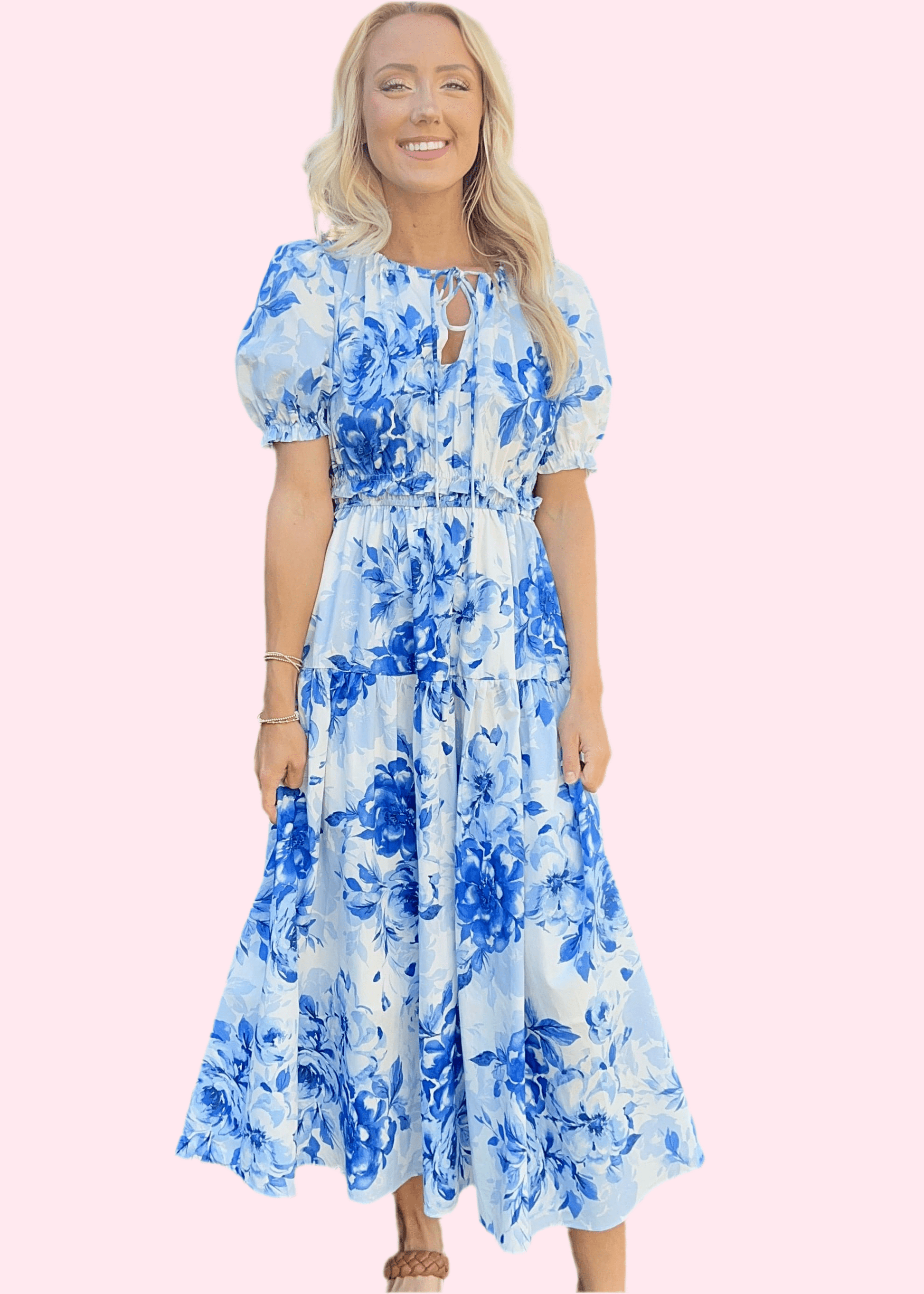 Full-length image of model in a blue floral tiered midi dress, smiling in front of pink background.