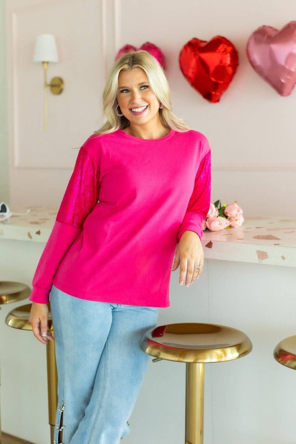Woman wearing a bright pink sweater and light blue jeans in a room with heart-shaped balloons.
