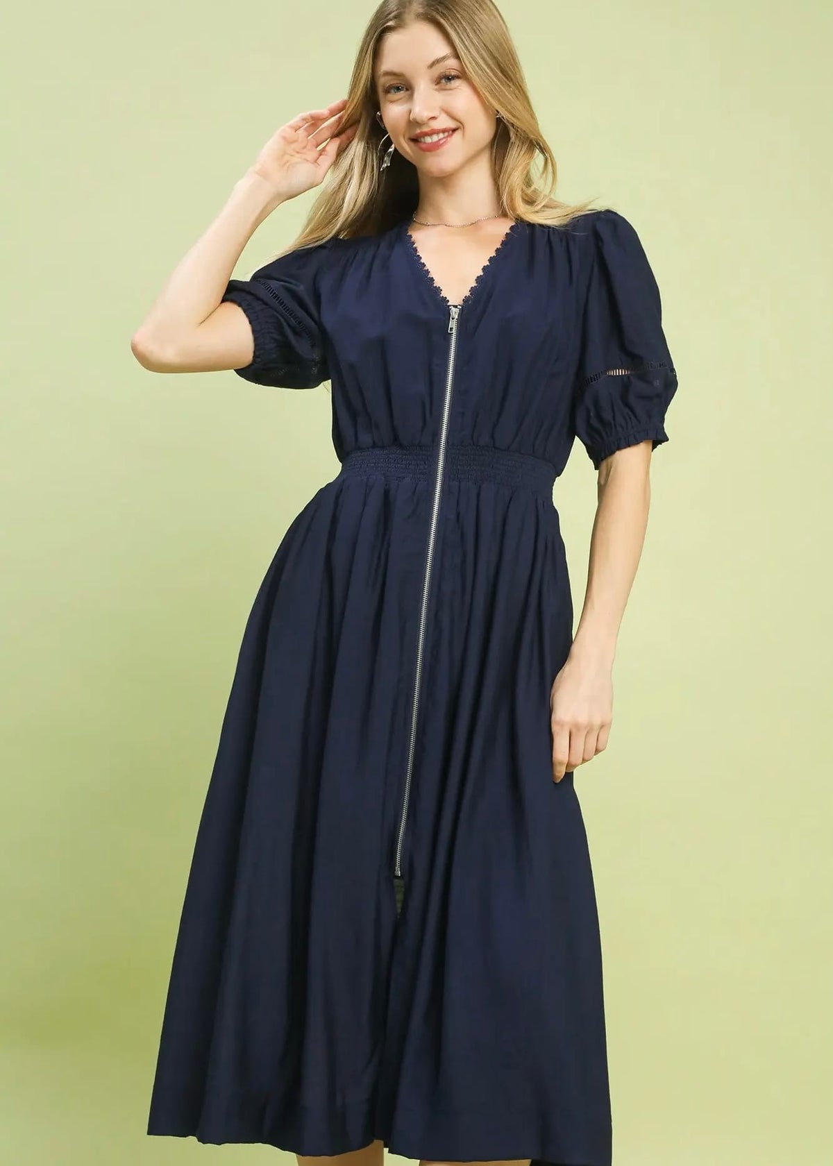 A woman modeling a navy blue midi dress with puff sleeves, a V-neckline, and a front zipper, set against a solid light green background.