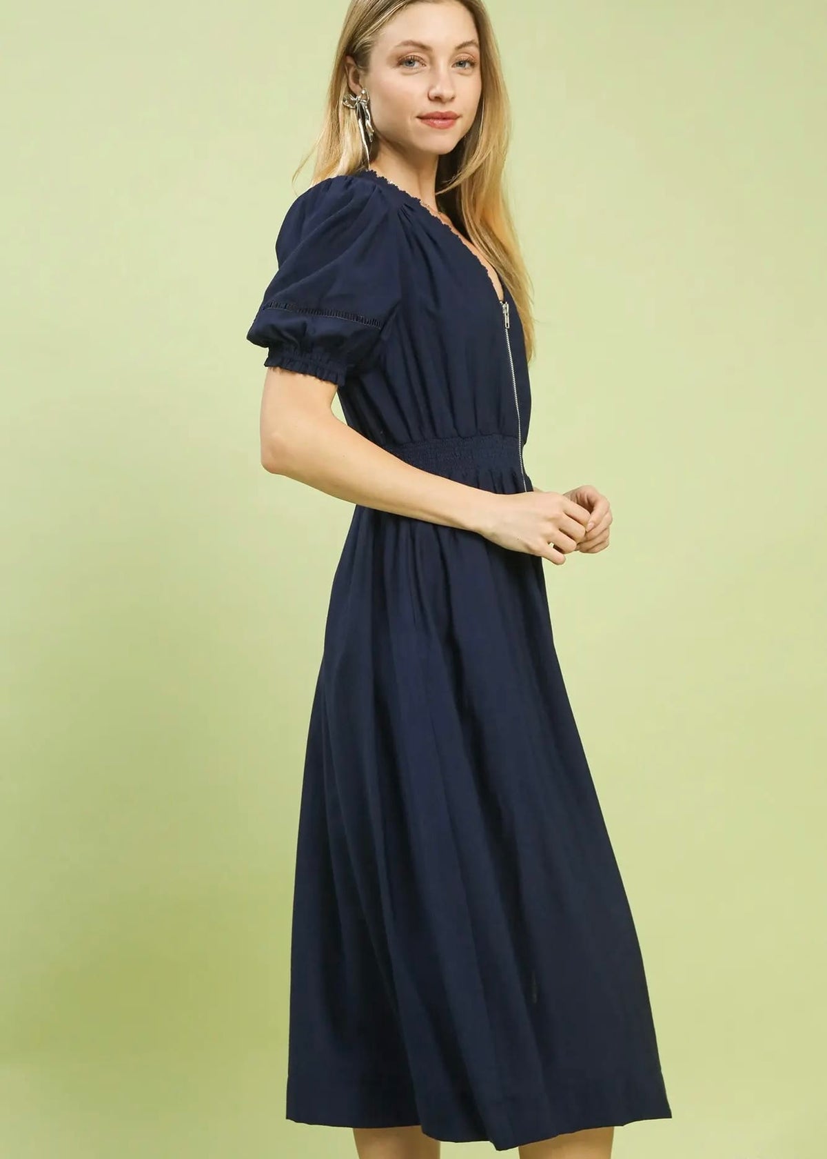 A woman modeling a navy blue midi dress with puff sleeves, a V-neckline, and a gathered waist, set against a solid pale green background.